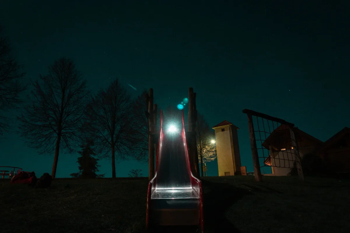 Night view of a playground with a slide, trees, and small buildings, illuminated by distant park lights. Parham Motamedifard