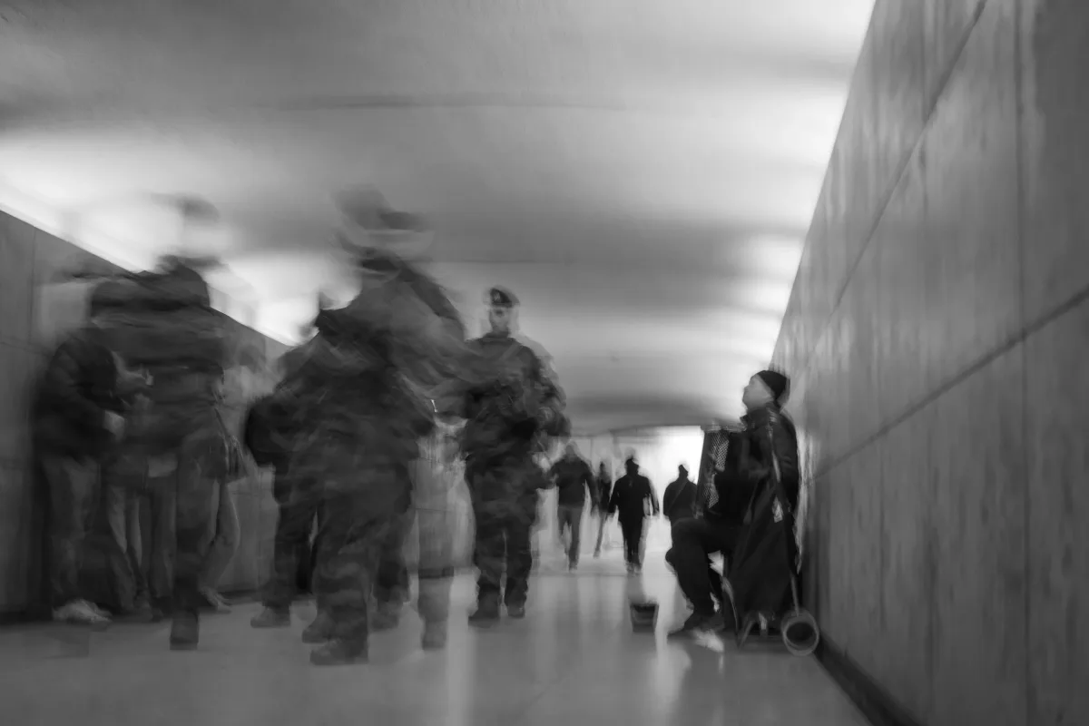 A black and white photo of a group of people walking through a tunnel. A musician is sitting against the wall, playing an accordion, wearing a hat and a coat. Parham Motamedifard