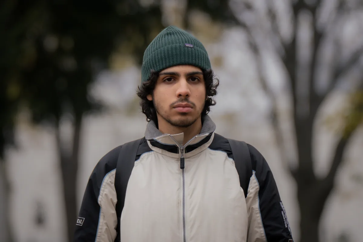 Young man with dark curly hair wearing a green beanie, beige and black jacket, and backpack, standing outdoors with a blurred background of trees.