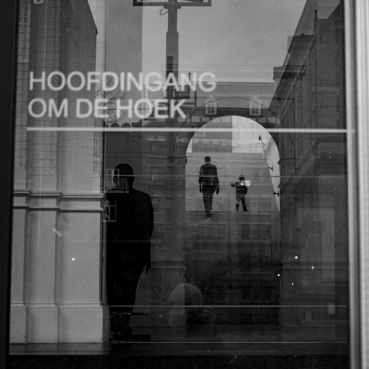 Reflection of a person walking down stairs with two children in the background, seen through a glass door with Dutch words 'HOOFDINGANG OM DE HOEK' written on it. Parham Motamedifard